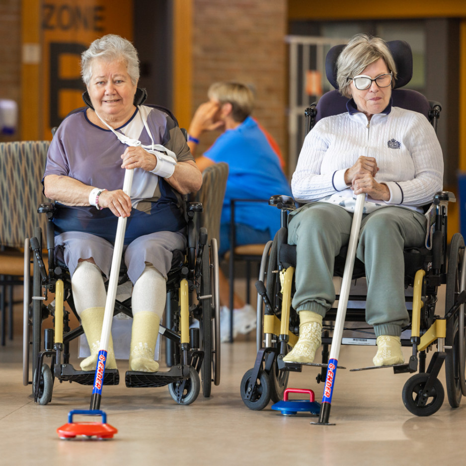 Two elderly women in wheelchairs play curling indoors using assistive devices